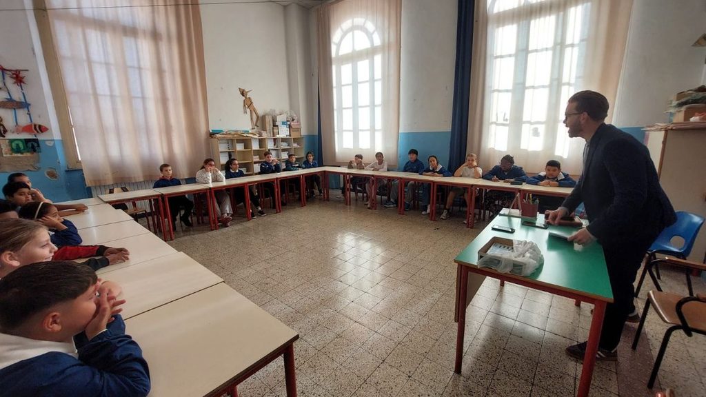 Alunni seduti ai tavoli di un'aula scolastica durante un incontro con un adulto in piedi