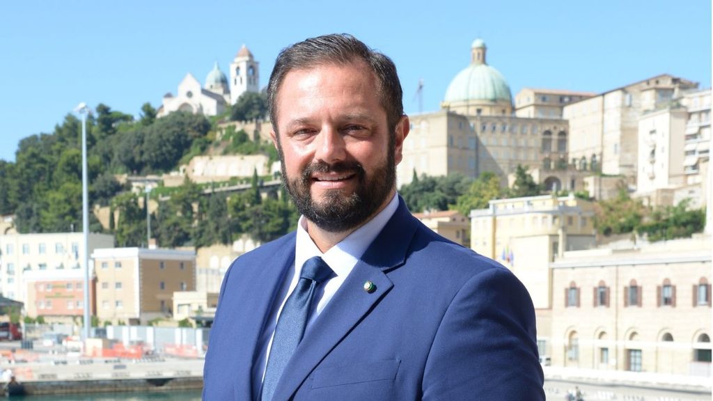 Uomo in abito blu davanti a uno scalo portuale con edifici sullo sfondo