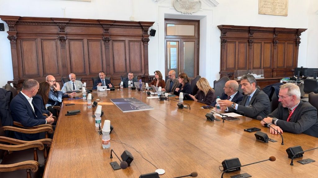 Partecipanti a una riunione istituzionale seduti attorno a un tavolo in una sala con pannelli in legno e microfoni.