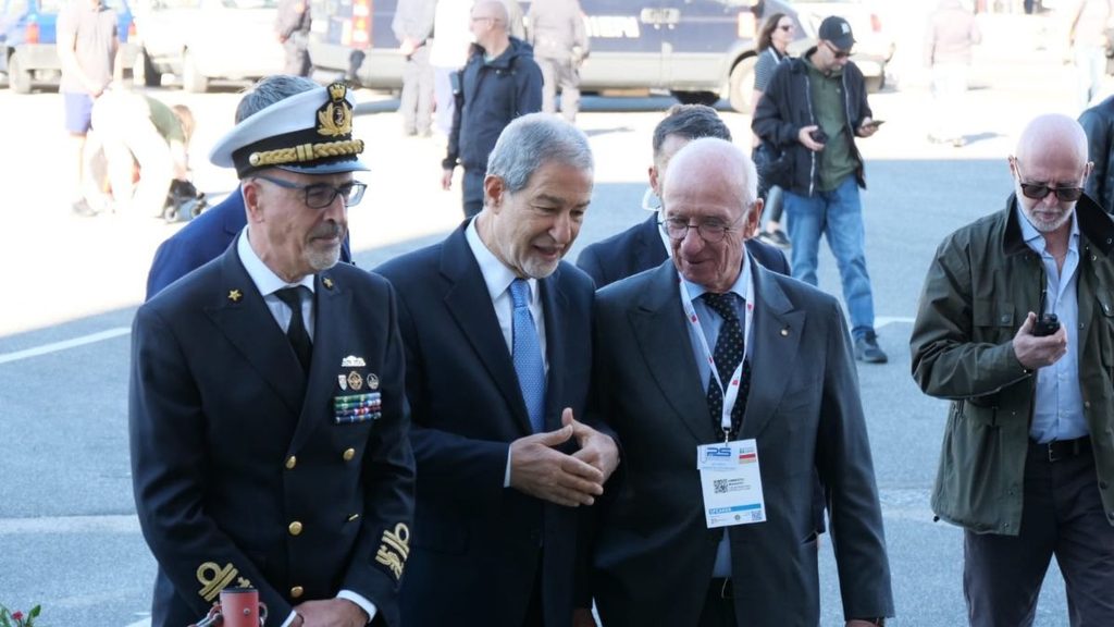 Tre uomini in primo piano, tra cui un ufficiale in uniforme della Marina, durante un evento all'aperto a Napoli.